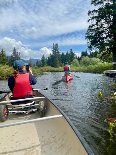 A picture taken from the stern of a canoe, while paddling down a narrow, calm river. The woman in the bow is taking a picture while a man paddles another canoe in the background.
